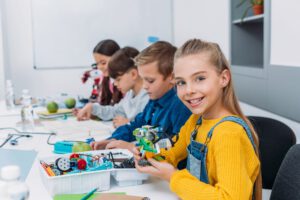 Lernen Vier Kinder sitzen an einem Tische und bauen technische Geräte aus Lego auf. Ein Mädchen im Vordergrund lächelt in die Kamera